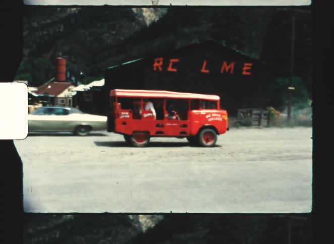Vintage red bus travels dirt road past dark building with orange letters and mountains in background