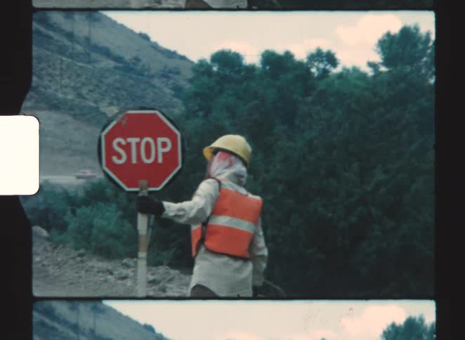 A construction worker wearing a hard hat safety vest and a veil is holding a stop sign near a wooded hillside
