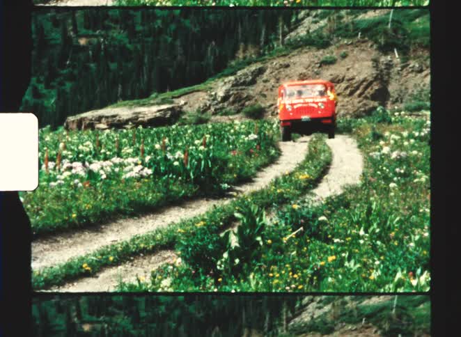 Red vehicle with passengers driving along narrow trail in green mountainous area