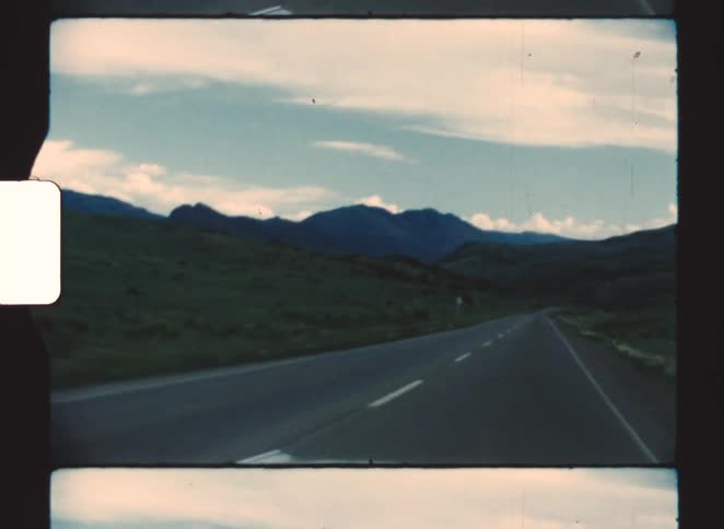 A car driving along a countryside freeway with green vegetation lining the road and a mountain range visible in the distance in the background