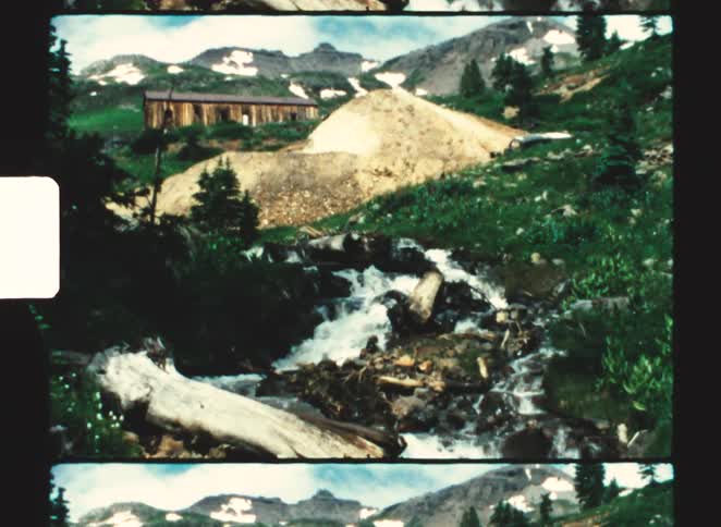 Snow dabbed mountains tower over a building and a sloping stream on a cloudy day