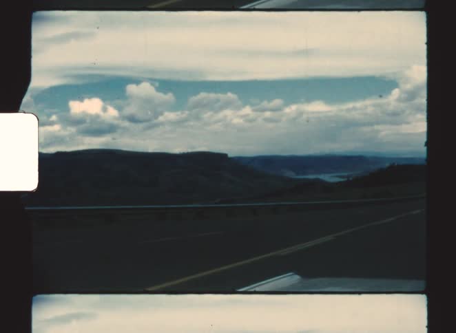 The perspective of a moving vehicle on a high mountain road captures a scenic view of a wide valley with a dark range of hills bordering a body of water under a dramatic blue and white cloudy sky