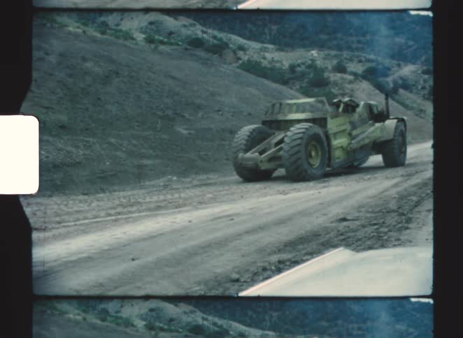 A large heavy duty faded green earth moving machine drives down a dirt road in a barren hilly landscape in vintage film