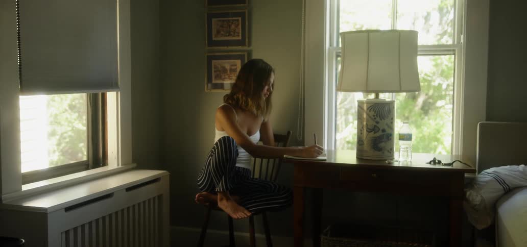 A woman sits by a table in a house writing on a notepad