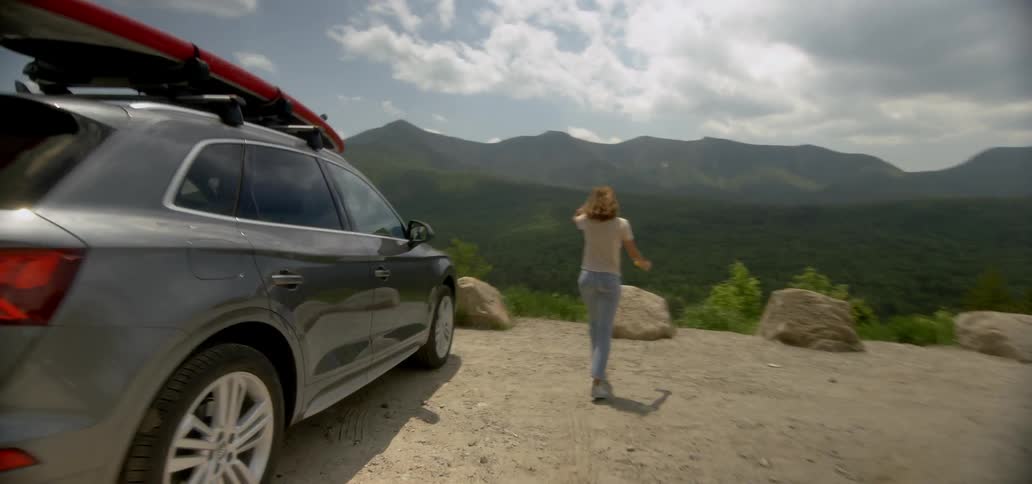A woman exits a car while parked in the mountains and climbs on a boulder