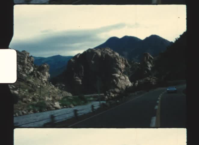 Car drives along winding road between rocky mountains and body of water under partly cloudy sky
