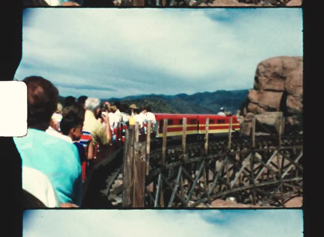 A miniature novelty train carrying passengers travels across a wooden trestle bridge set against a mountain landscape under a bright sky