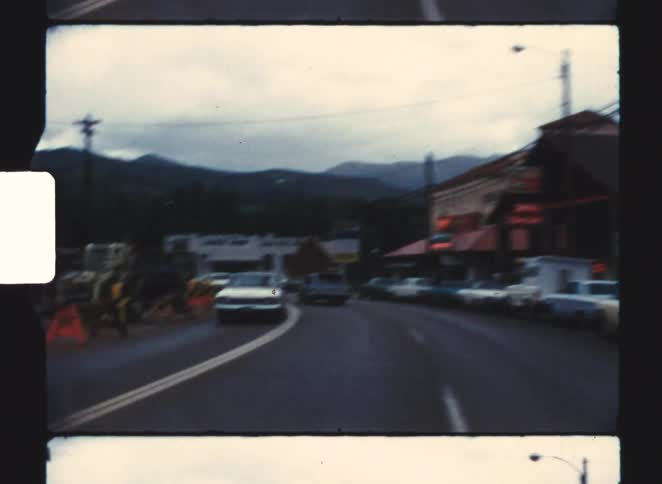 Curved street runs through commercial area with parked cars and mountains under cloudy sky