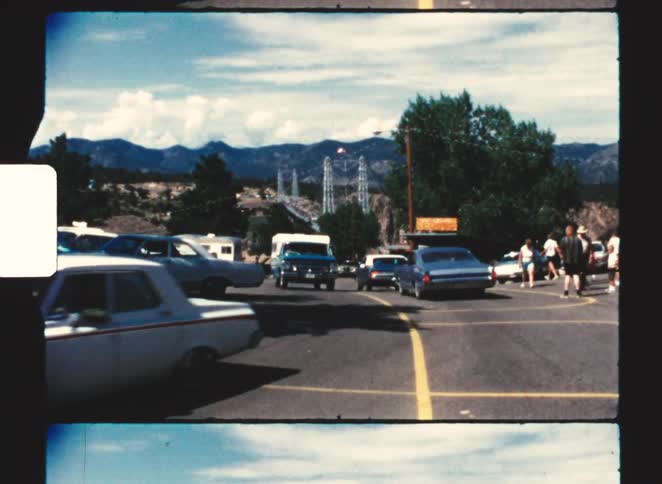 A busy street at midday with cars driving past a group of people gathered on the sidewalk and several cars parked along the roadside