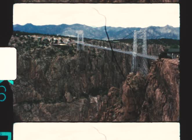 Vintage footage captures a distant view of the iconic royal gorge bridge suspended precariously over a massive rugged canyon with a mountain range stretching across the horizon
