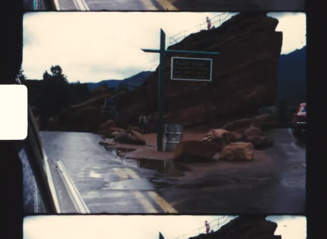 A large distinctive reddish rock formation sits beside a wet divided highway marked by a crossshaped post