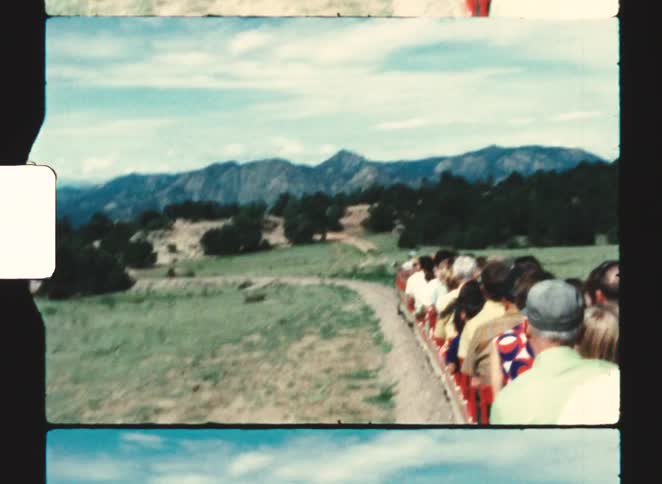 A happy young boy wearing a red shirt rides a small train with other passengers on a track surrounded by green hills and distant mountains under a blue sky