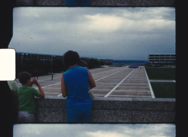 A mother and her two sons standing on a rooftop watching a grass field at midday under a cloudy sky