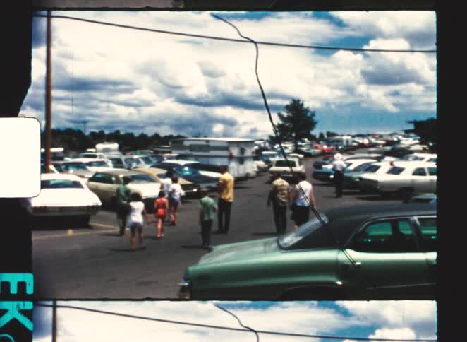 A large outdoor car park filled with cars with a big crowd of people walking around at midday