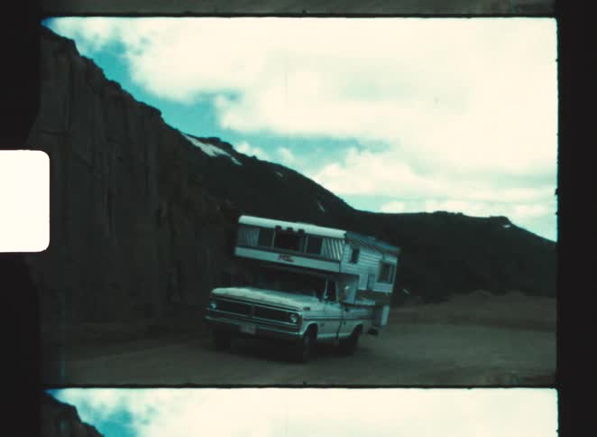 A camper truck drives along a dirt road through a rocky mountain landscape