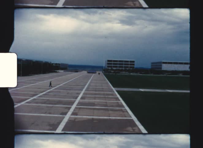A lone person stands on a vast geometrically marked concrete plaza between modernist buildings under a dramatic cloudy sky creating a monumental cold perspective
