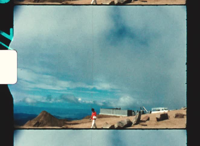 Person wearing red clothing walks across an open area with structures and a mountainous horizon in the background