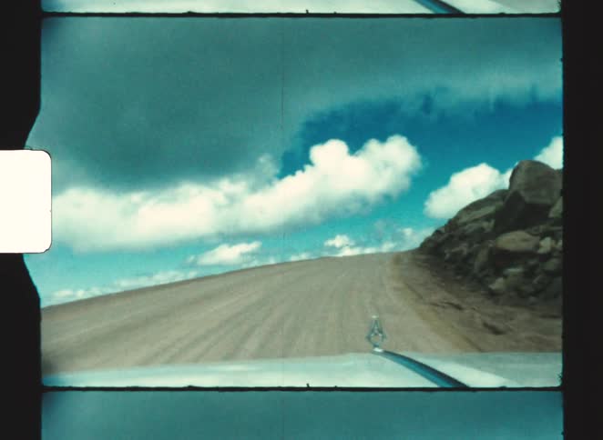 pikes peak colorado springs 1970  From the dashboard of a classic car the view shows a dirt road winding up a steep mountainside under a dramatic cloudy sky