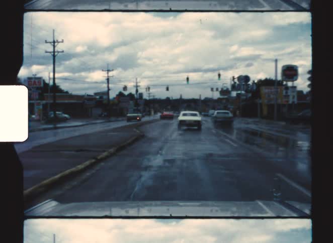 Wet street with moving vehicles under cloudy sky and visible traffic lights ahead
