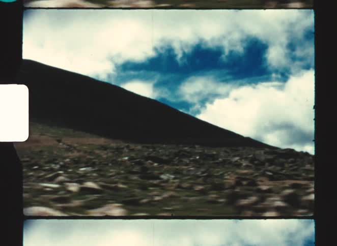 A sloping dark hillside rises from rocky ground in the foreground set beneath a sky filled with layered white and blue clouds