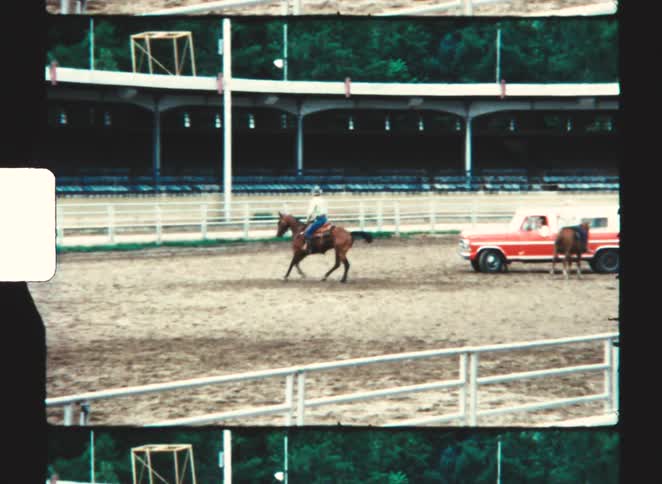 A man in a cowboy hat rides a brown horse in a dirt arena passing a red pickup truck parked near a white fence with stadium seating visible behind