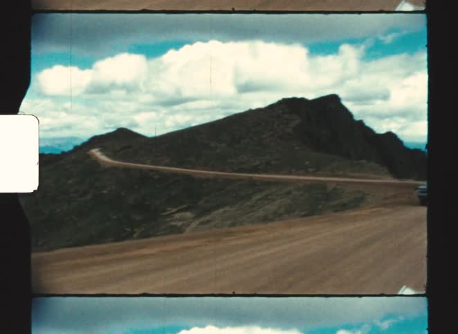 A vintage dark colored car drives quickly on a winding dirt road up a barren mountain, pikes peak under a cloudy blue sky in colorado springs