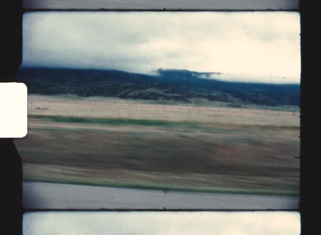 Captured from a moving vehicle the camera sweeps across a vast flat plain leading up to a rugged mountain range shrouded in thick lowhanging clouds