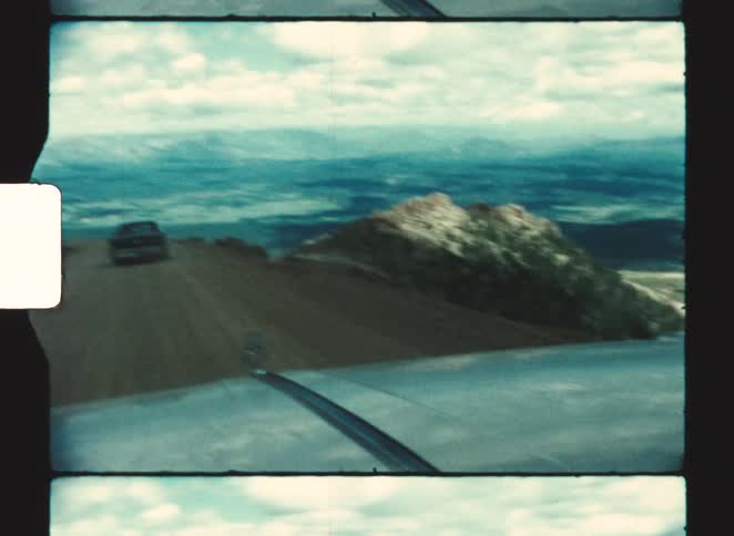 A drivers view from a vintage car travels down a dirt road with steep dry hillsides and distant plains visible under a cloudy sky