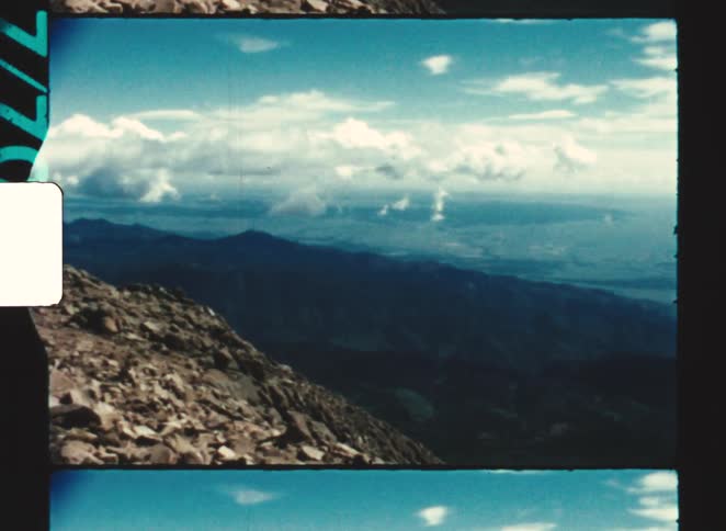 Wide view of a valley and plains seen from a rocky slope under a partly cloudy sky