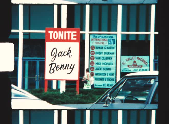 A buildings glass facade featuring two promotional signs one advertising a tonite jack benny show and another listing a 1970 broadmoor international theatre schedule with a partial view of a car and a few people in the foreground