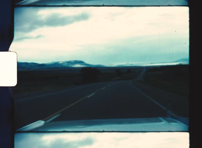 A car drives down an empty highway toward distant mountains under a cloudy sky