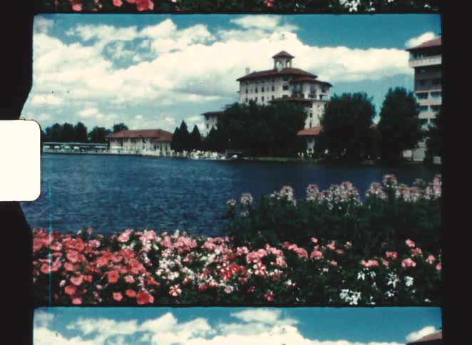 People sit in a boat in a lake bordered by flowers and apartment buildings on a cloudy day