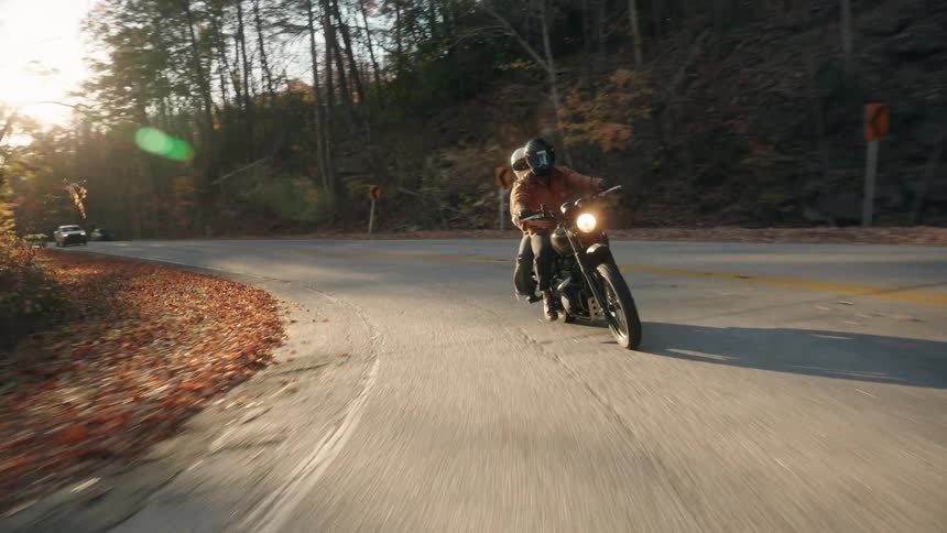 Motorcyclist riding along a curved paved road surrounded by forest with low sunlight
