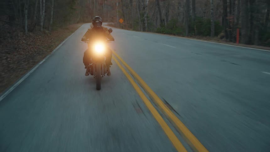 A person riding a motorcycle with a passenger seated behind both wearing helmets the bikes headlight on as they travel at sunset