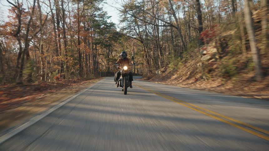 Two people riding a motorcycle in the countryside at midday in the fall both wearing helmets with a car following behind them and trees lining both sides of the road