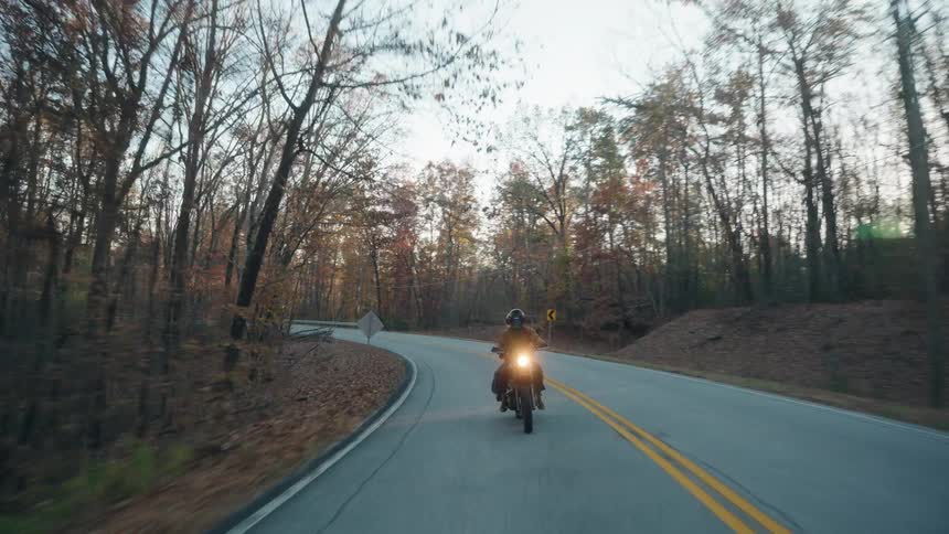 Motorcycle travels on a road lined with trees in fall colors as the sun sets in the distance