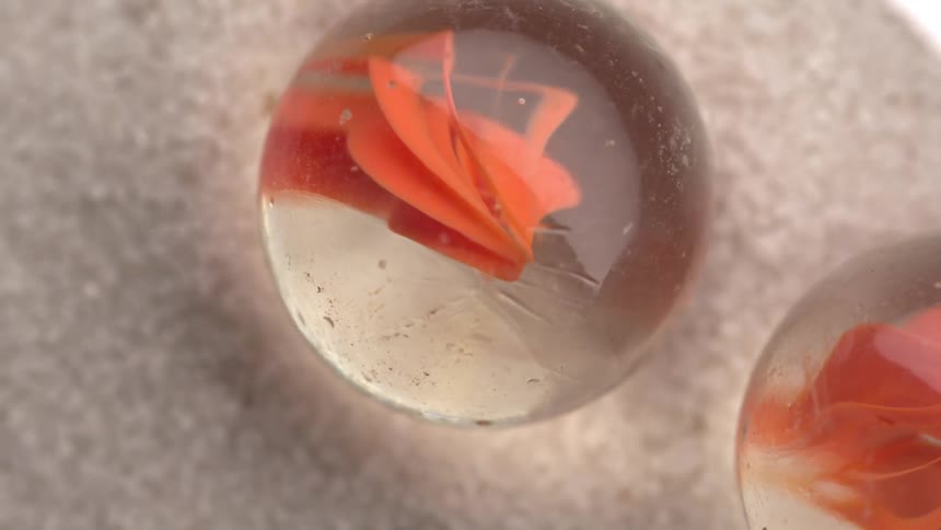 A clear glass marble with an internal orange spiral pattern rests on fine white sand next to a second marble