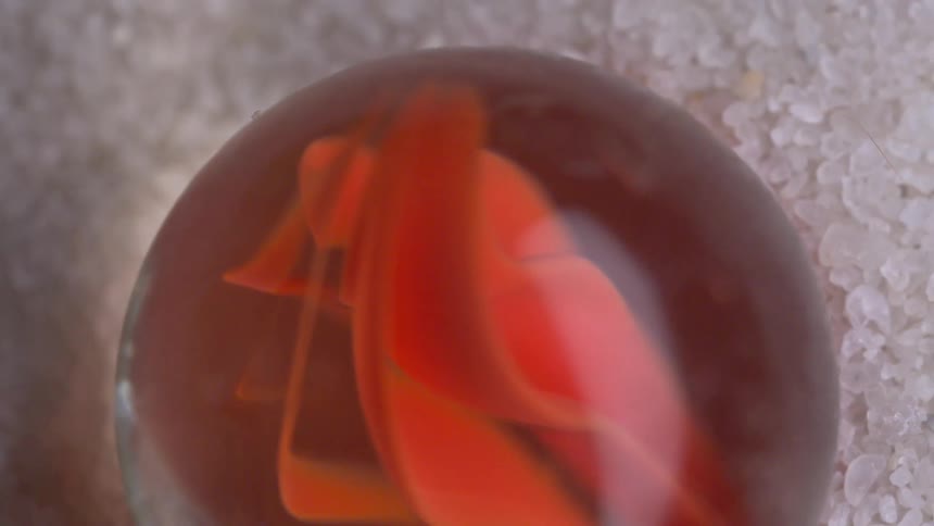 A macro shot pans slowly across a textured bed of coarse white crystals revealing a clear glass marble with a vibrant orange swirl suspended within its smooth surface
