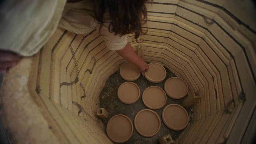 A potter reaches down carefully placing a small clay bowl among other unfired pieces at the bottom of a large kiln