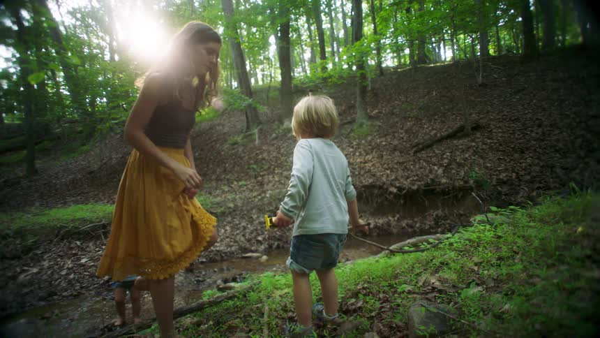 Male child on a grove carrying a toy in one hand and a branch on another kicks the floor and then walks to a young adult woman who inspects his feet while a female toddler stands on the background next to a stream of water outdoors during day time