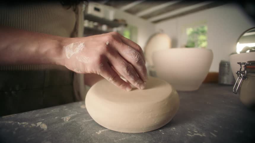 A blurred shot of an artisan in a pottery studio shifts focus to a closeup of their hands smoothing and detailing a freshly formed round piece of clay on a dusty workbench using a small sponge