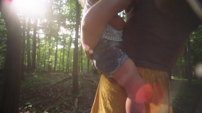 A woman wearing a dark top and a mustard yellow skirt carries a young child through a sun drenched forest