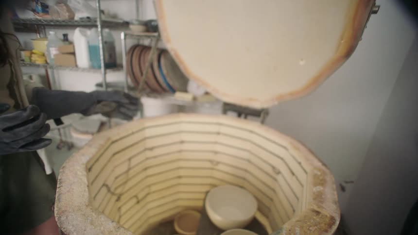 A woman opens a ceramic kiln and pulls a white bowl out of it