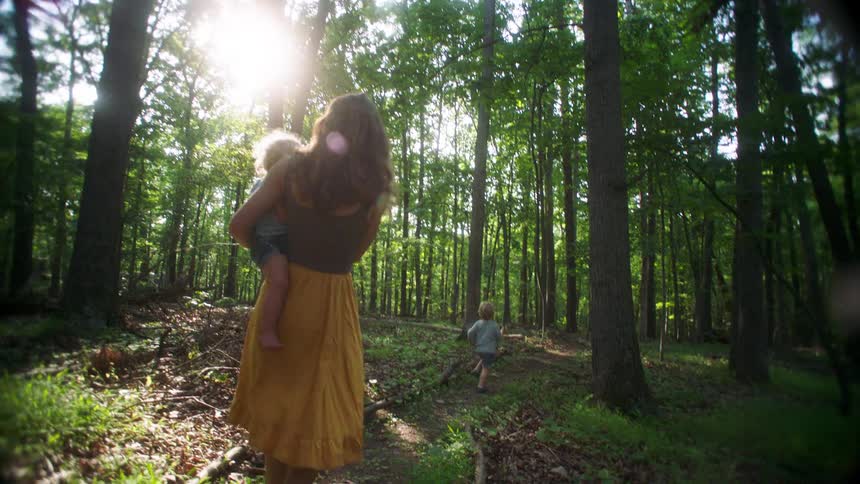 Woman walks along a forest path carrying a child while another walks ahead among the trees
