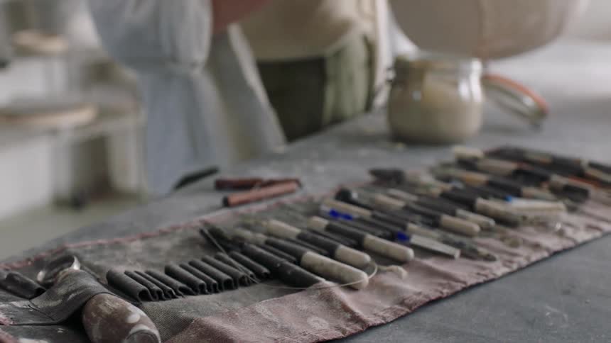Several paintbrushes are on top of an old cloth and a person behind them is rummaging through a jar