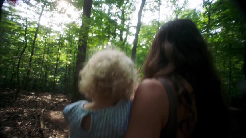 Woman carries a child while walking along a forest trail lit by sunlight