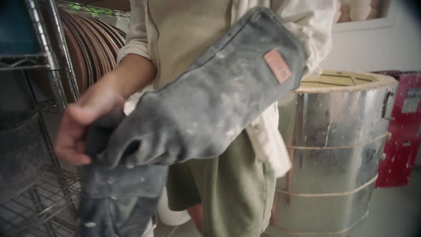 Young adult woman in a pottery studio putting on heavy duty gloves and then walking towards a tin container indoors during day time