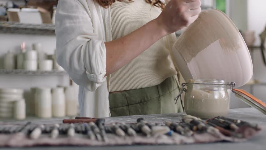 A woman in a white shirt applies a beige glaze to a large ceramic bowl using a thin paintbrush she works in a pottery studio with various tools and finished vessels visible on the shelves behind her