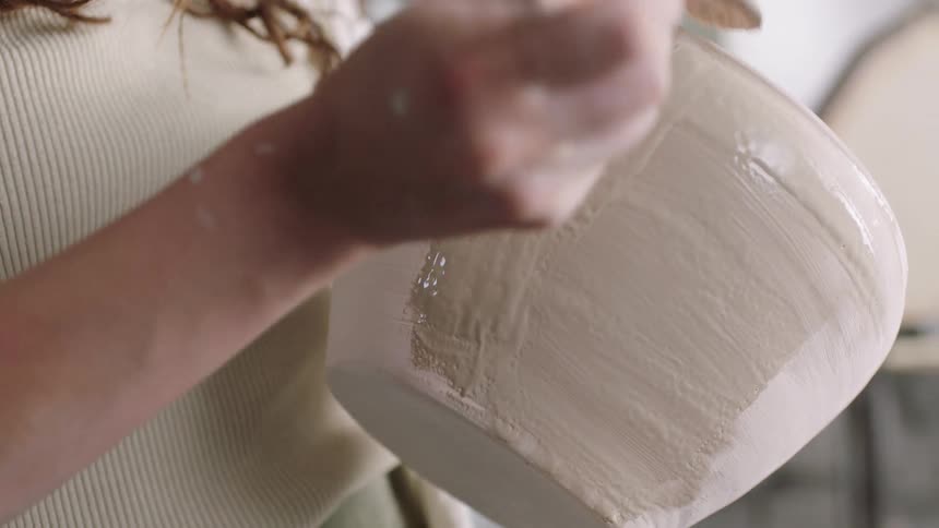 A woman holds a ceramic pot carefully applying a thick beige glaze with a small brush in smooth deliberate strokes