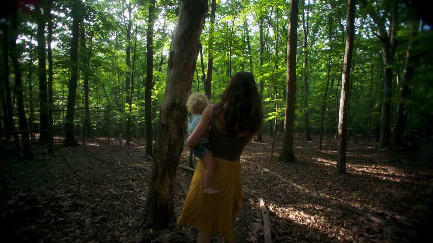 Young adult woman wearing a yellow skirt and a brown blouse carrying a female toddler on her arms through a forest outdoors during day time